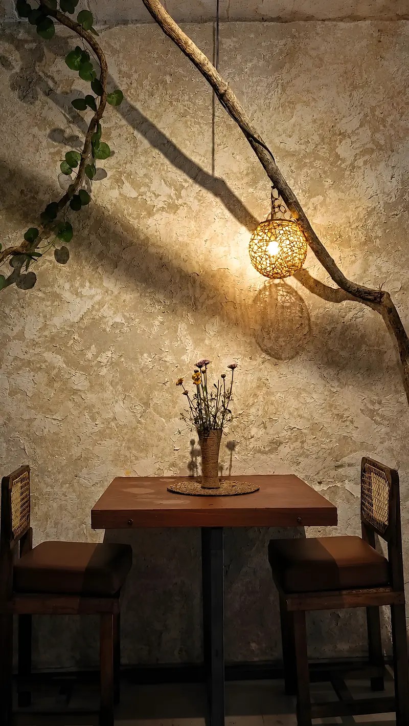 A table for two beneath a woven pendant lamp and a branch of vines against a plaster wall